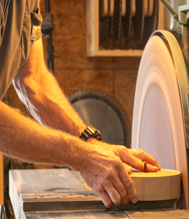 Hands sanding a round piece of cygnus lumber on a disk sander in a workshop.
