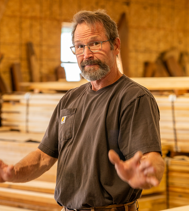 Man in glasses gesturing in a Cygnus lumber warehouse, surrounded by stacked wood planks.