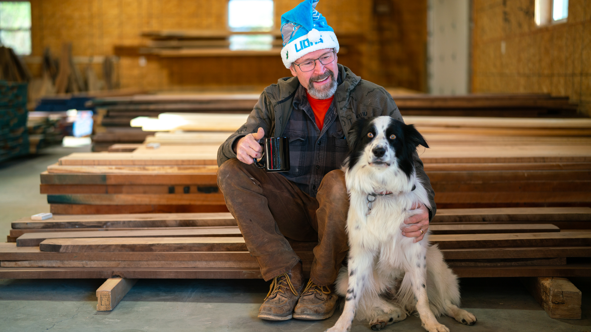 Woodworker with a coffee mug sitting beside his dog near stacked lumber.